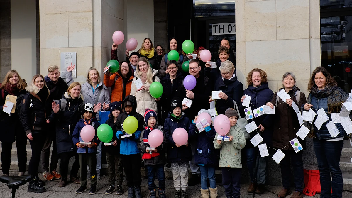 Gruppenfoto der Beteiligten auf einer Treppe - viele halten Luftballons in rosa, grün und lila in den Händen