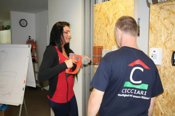 Foto einer Frau mit Bohrmaschine. Sie übt mit einem Ziegelstein, der an der Wand befestigt ist. Ein Jugendlicher sieht zu.